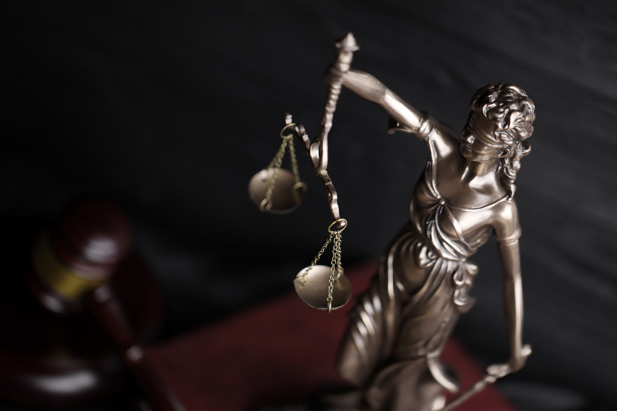 The Statue of Justice blindefolded and holding scales on a stack of brown books with judge's gavel in the background.
